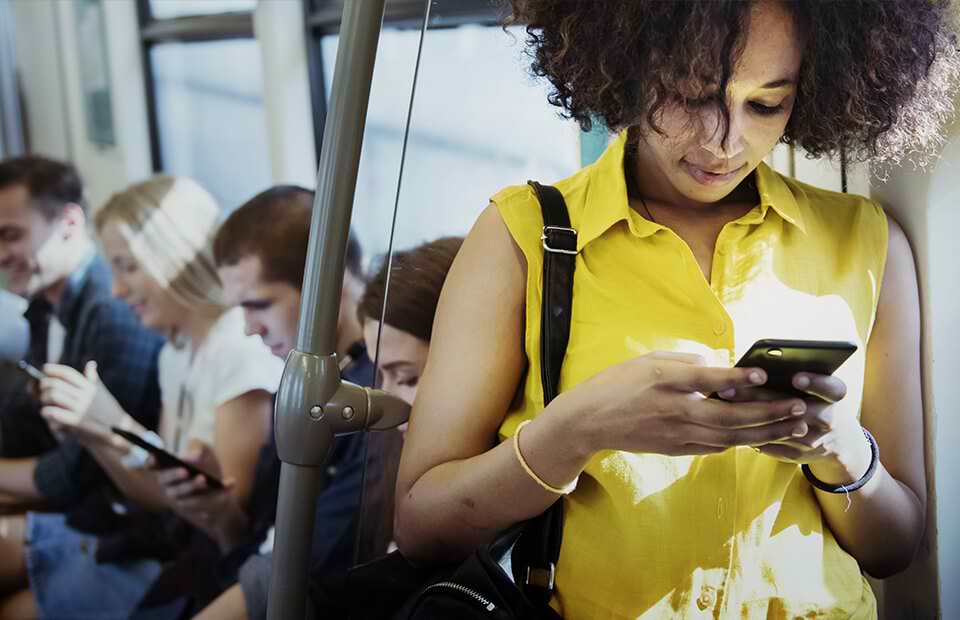 Junge Frau stehend mit Handy im Zug, hinter ihr sitzend weitere Fahrgäste mit digitalen Endgeräten