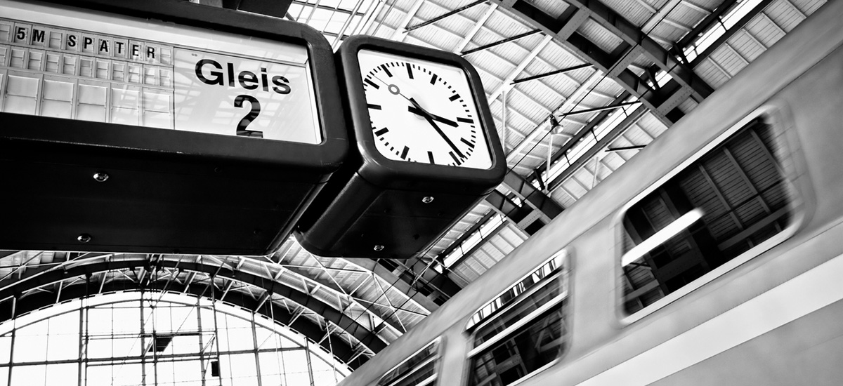 Zug fährt mit 5 min Verspätung - sichtbar auf der Anzeigentafel neben der Uhr - in den Bahnhof ein