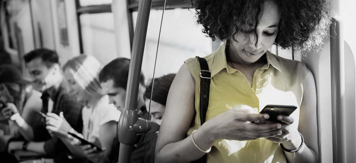 Young woman standing next to a train door is reading on her mobile phone. In the background, other young people are sitting on their seats along the train's window.