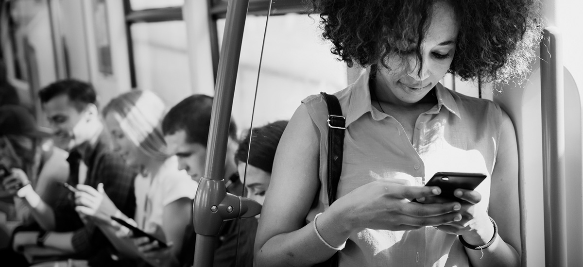 Junge Frau lehnt Handy-lesend neben der Zugtür. Im Hintergrund sitzen weitere junge Menschen mit ihren mobilen Geräten auf Sitzen entlang der Fensterfront des Zuges.